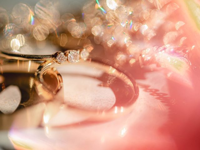 Detailný záber na dva zlaté prstene, jeden s tromi malými diamantmi, obklopené iskrivými farebnými bokeh svetlami a jemnou, zasnenou ružovou žiarou v pozadí.