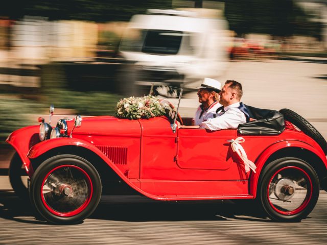 Dvaja muži vo formálnom oblečení riadia historický červený kabriolet ozdobený kvetmi a bielymi stuhami a uháňajú po ulici s rozmazaným pozadím.