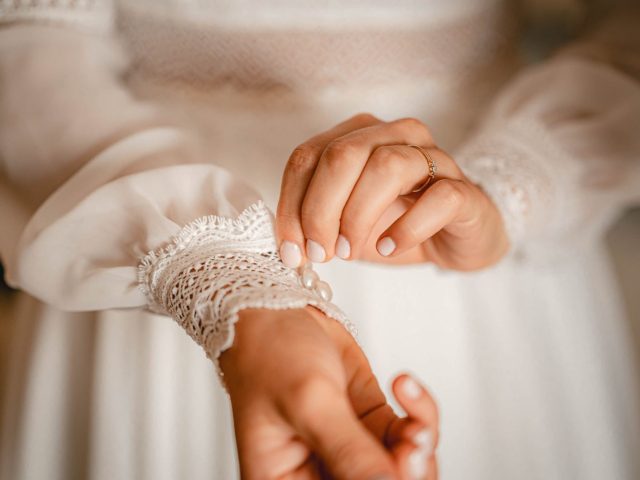 Osoba si zapína jemnú čipkovú manžetu bielych šiat s dlhými rukávmi, pričom sa zameriava na svoje ruky a zložité detaily látky.