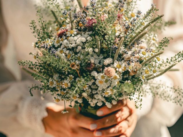 Nevesta v bielych šatách drží kyticu z poľných kvetov a zelene s jemnými bielymi, fialovými a žltými kvetmi v jasnom prirodzenom slnečnom svetle. Nechty má nalakované na bledoružovo.