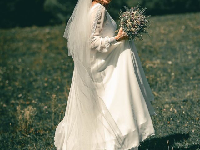Nevesta v bielych šatách a závoji s kyticou kvetov stojí vonku na trávnatom poli so stromami a kopcami v pozadí, usmieva sa a pozerá sa cez plece.