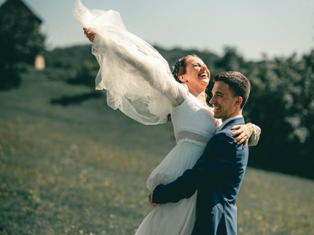 Ženích v modrom obleku zdvíha nevestu v bielych šatách, obaja sa radostne usmievajú na trávnatom kopci pod jasnou oblohou. Nevesta má zdvihnuté ruky a závoj za ňou splýva.