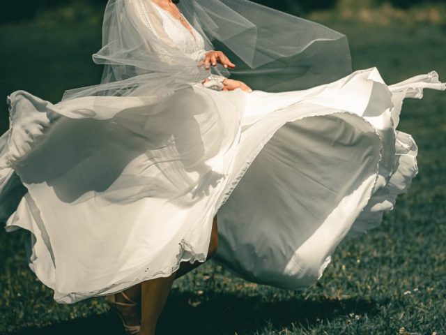 Nevesta v splývavých bielych šatách a závoji sa radostne krúti na trávnatom poli a jej šaty sa v slnečnom svetle vlnia okolo nej.