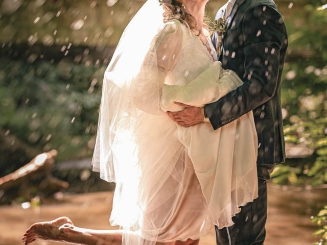 Nevesta a ženích stoja v plytkej vode a radostne sa bozkávajú. Nevestu v bielych šatách a závoji dvíha ženích, ktorý má na sebe oblek s vyhrnutými nohavicami. Slnečné svetlo a kvapky vody vytvárajú čarovnú, romantickú atmosféru.