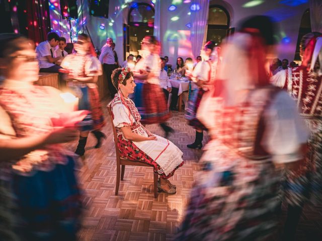 Žena v tradičnom ľudovom odeve sedí na stoličke uprostred tanečného parketu, obklopená rozmazanými tanečníkmi v pestrofarebných krojoch, so slávnostnými svetlami a divákmi v pozadí.