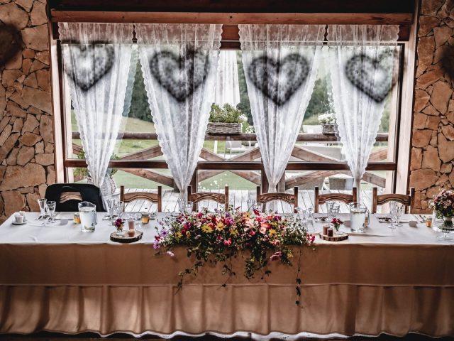 Zdobený svadobný stôl s bielym obrusom, kvetinovou výzdobou a elegantným prestieraním sa nachádza pred veľkými oknami s čipkovanými závesmi a zavesenými vencami v tvare srdca. Scénu rámujú rustikálne kamenné steny.