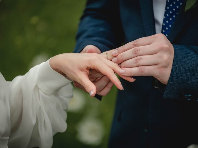 Detailný záber na osobu v modrom obleku, ktorá navlieka prsteň na prst inej osoby, obe stoja vonku na rozmazanom zelenom pozadí.