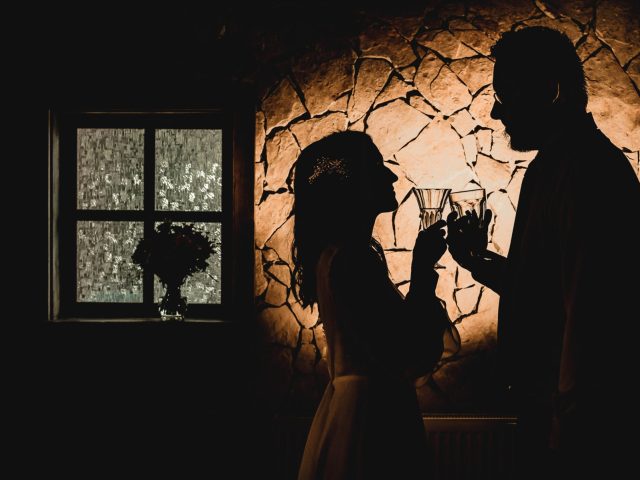 Silueta dvojice, ktorá si štrngá pohármi s vínom pred teplým osvetlením kamennej steny, v pozadí vidno okno a vázu s kvetmi.
