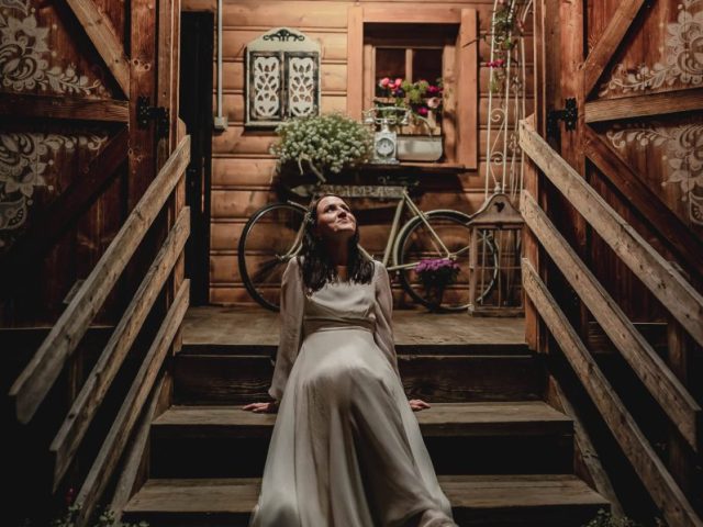 Žena v dlhých bielych šatách sedí v noci na drevených schodoch, za ňou sú otvorené rustikálne dvere a teplo osvetlený drevený dom. Verandu zdobí bicykel s košíkom a kvetmi.