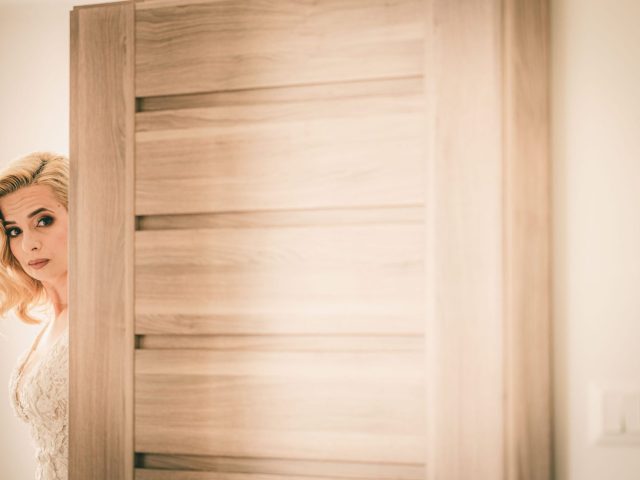 Za mierne pootvorenými drevenými dverami stojí čiastočne viditeľná žena so svetlými vlasmi a bielymi šatami a zamyslene hľadí von. Scéna je jemne osvetlená a v teplých tónoch.