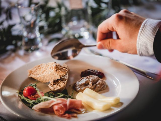 Osoba drží lyžicu nad tanierom s chlebom, paštétou, syrovými tyčinkami, prosciuttom, zeleninou a cherry paradajkou, položeným na bielom obruse.