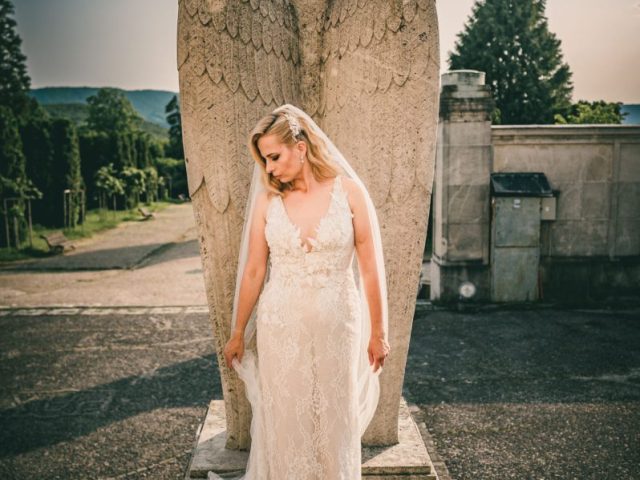 Nevesta v čipkovaných svadobných šatách stojí pred veľkou kamennou sochou anjela v exteriéri, v pozadí ju osvetľuje slnečné svetlo a vidno zeleň.