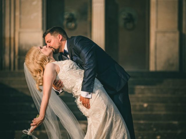 Ženích v tmavom obleku sa ponorí a pobozká svoju nevestu, ktorá má na sebe biele čipkované šaty a závoj, keď stoja na schodoch pred veľkolepou budovou. Nevesta drží kyticu bielych kvetov a obaja vyzerajú radostne.