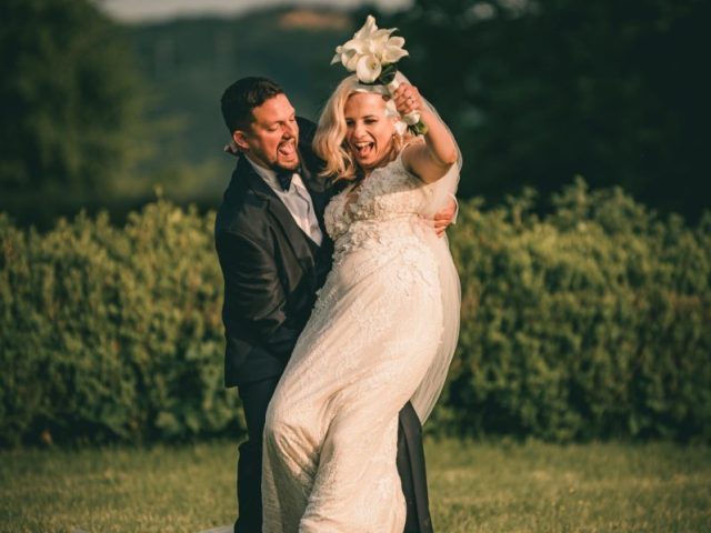 Ženích v tmavom obleku radostne dvíha svoju usmievavú nevestu v bielych čipkovaných šatách, ktorá drží kyticu. Stoja na trávnatom trávniku so stromami a kríkmi v pozadí.